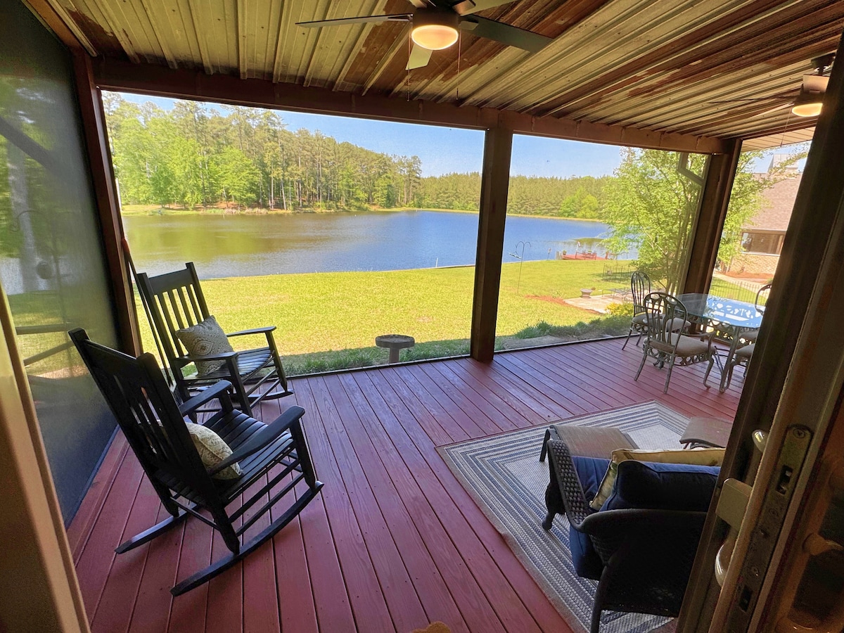 A screened porch features two rockers and a small table, overlooking a serene fishing lake. A plush seating area with a rug is visible, while lush green grass meets the water's edge, framed by trees in the background, creating a peaceful retreat.