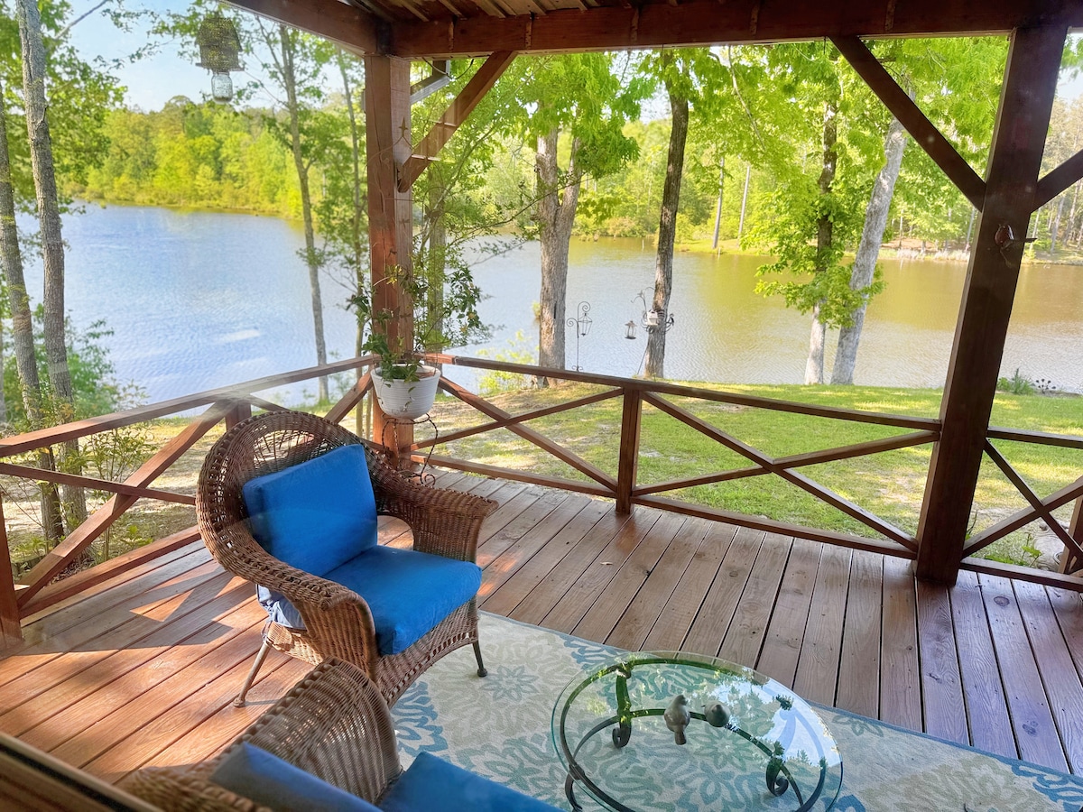 A shaded porch is showcased, featuring a blue cushioned chair and a glass table. Lush greenery surrounds the area, and a tranquil lake can be seen in the background, reflecting the peaceful environment. Soft natural light illuminates the wooden deck.