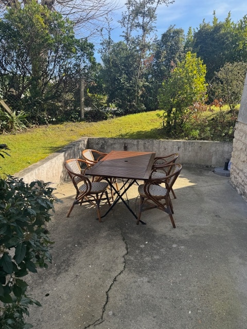 A private terrace is shown, featuring a wooden table surrounded by four wicker chairs. Lush greenery and shrubs are visible in the background, creating a serene outdoor space. The concrete floor is well-maintained, providing an inviting area for relaxation.
