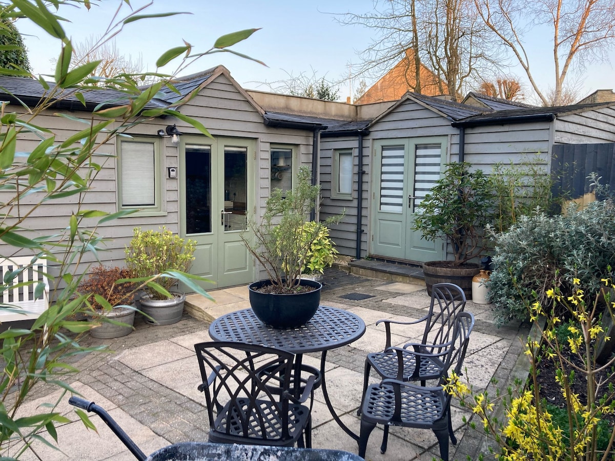 A private patio area is presented with gray stone paving, featuring a round table with four matching chairs. Potted plants and greenery surround the outdoor space, while muted green doors lead into the cabin. Soft natural light enhances the inviting atmosphere.