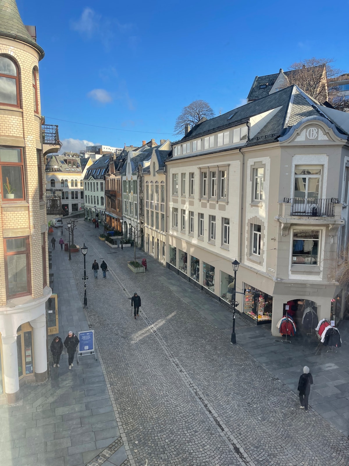 A quiet cobblestone street is depicted, lined with historic buildings showcasing various architectural styles. The scene includes pedestrians strolling casually, with sunlight illuminating the facades. Storefronts, adorned with displays, add to the vibrant atmosphere of this central shopping area.