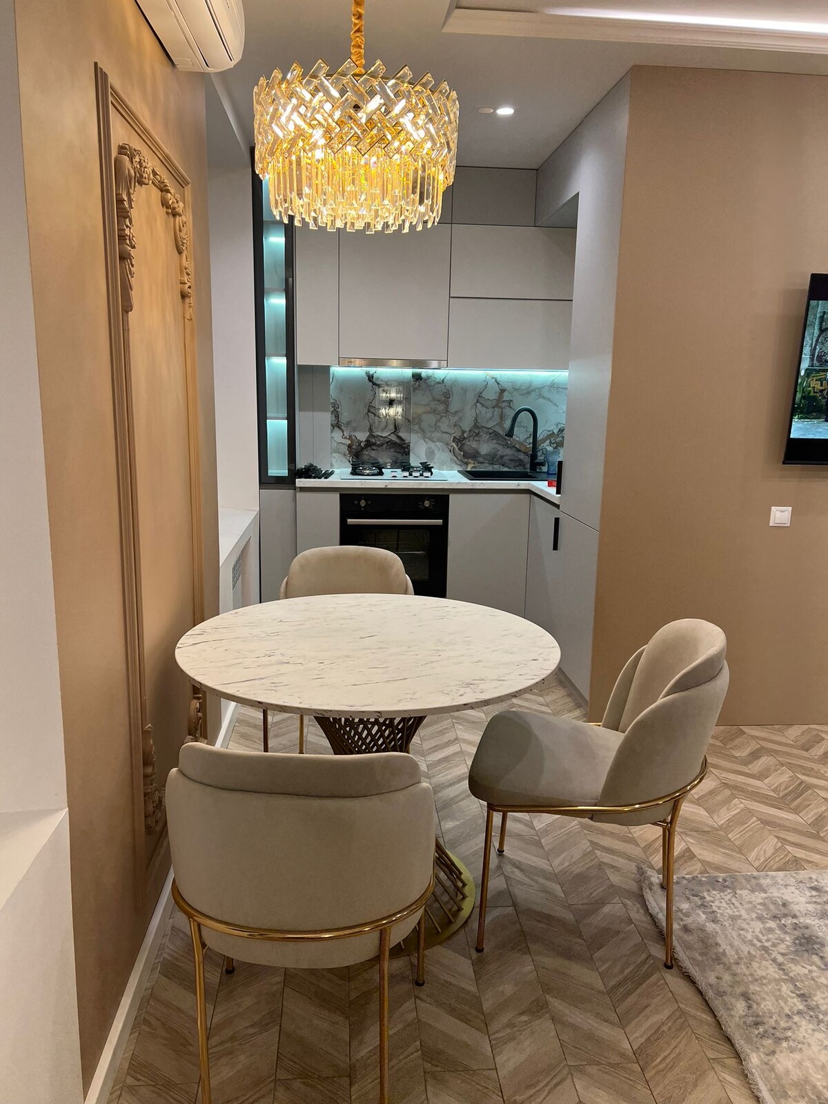 A modern dining area features a round marble table surrounded by three upholstered chairs with gold accents. A stylish chandelier hangs above, illuminating the space, while the kitchen is visible in the background with sleek cabinetry and a marble backsplash.