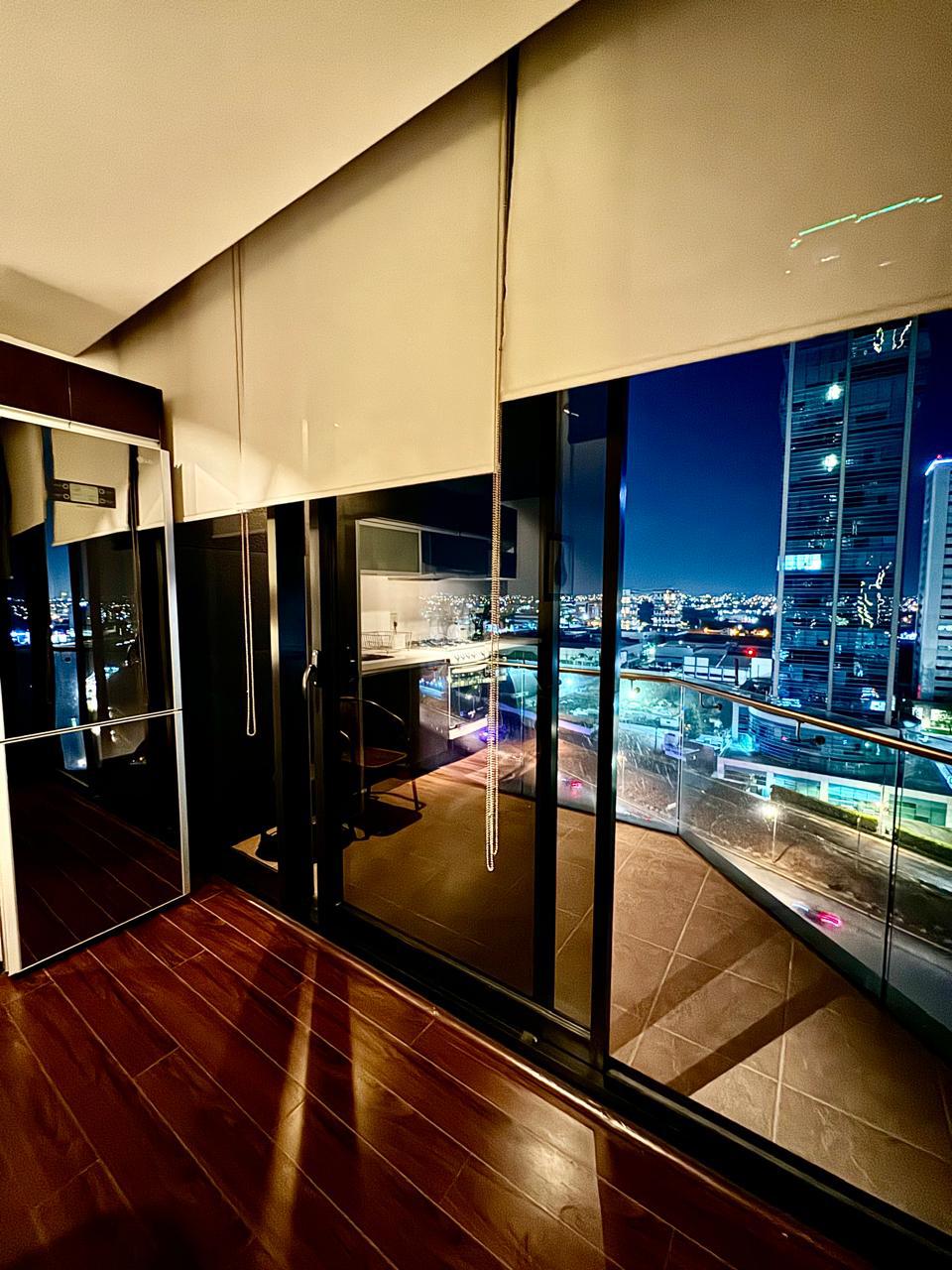 A modern loft interior is depicted, featuring floor-to-ceiling windows that offer a sweeping view of the city skyline at night. The warm wood flooring contrasts with the sleek, dark cabinetry. Soft lighting enhances the welcoming ambiance, creating a serene space for relaxation.