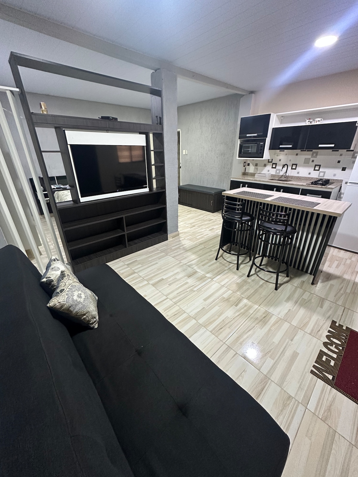 An open-concept living area is shown, featuring a sleek black sofa alongside a modern kitchenette equipped with dark cabinetry and stainless steel appliances. A flat-screen television is mounted on a pedestal, and three black bar stools are situated at the kitchen counter, creating an inviting space.