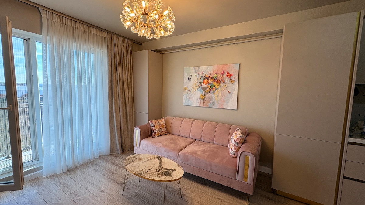A light-filled living area features a soft pink sofa adorned with decorative pillows. An elegant chandelier hangs overhead, and an abstract wall art piece adds color to the space. Sliding glass doors open to a balcony, offering outdoor access and scenic views.