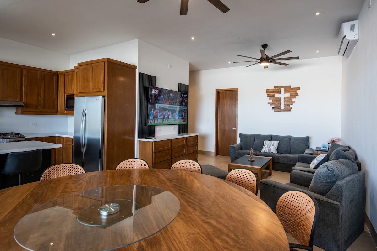 A spacious open-plan living area features a round wooden dining table with a glass top and several wicker chairs. A comfortable seating arrangement with gray sofas surrounds a coffee table. A large wall-mounted TV is visible, complemented by warm wooden cabinetry.