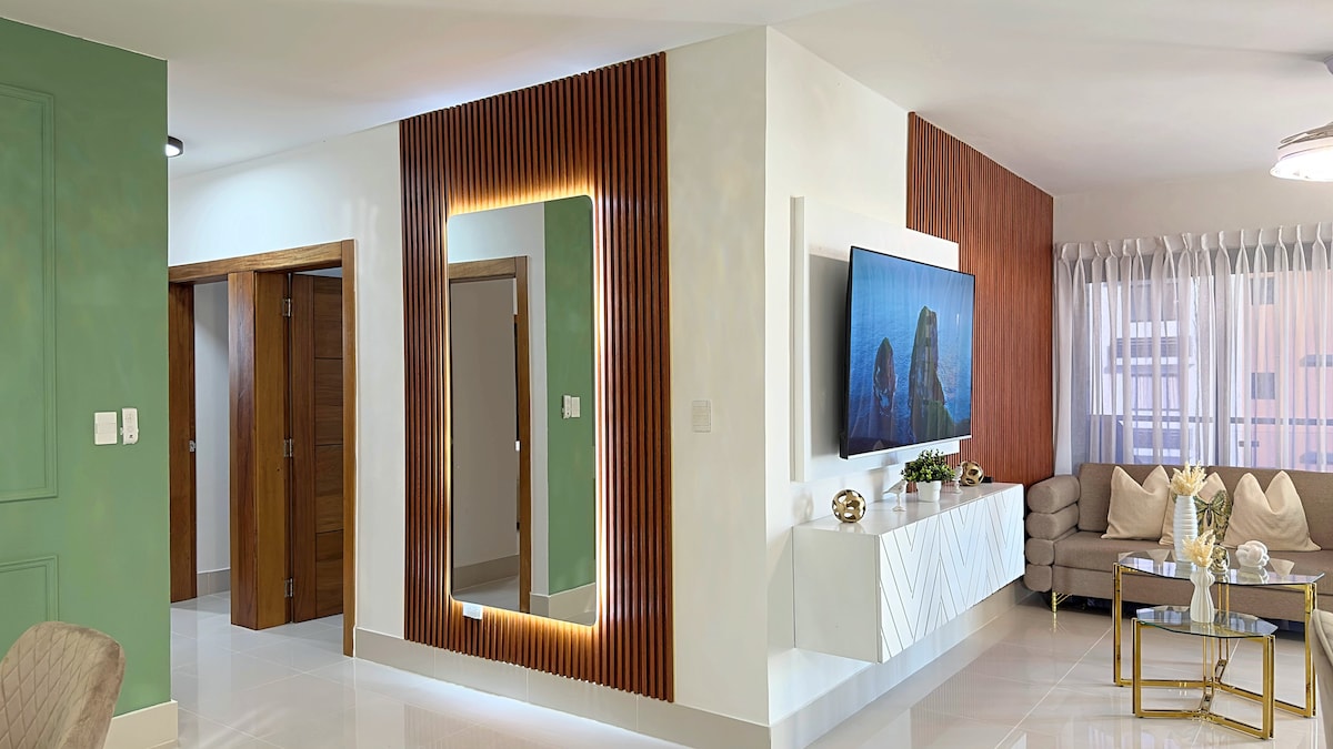 An open living space is featured, showcasing a large mirror framed with warm lighting. The area includes a modern television, a gray sectional sofa, and a white side table with decorative items. Natural light flows in through sheer window treatments, enhancing the bright atmosphere.