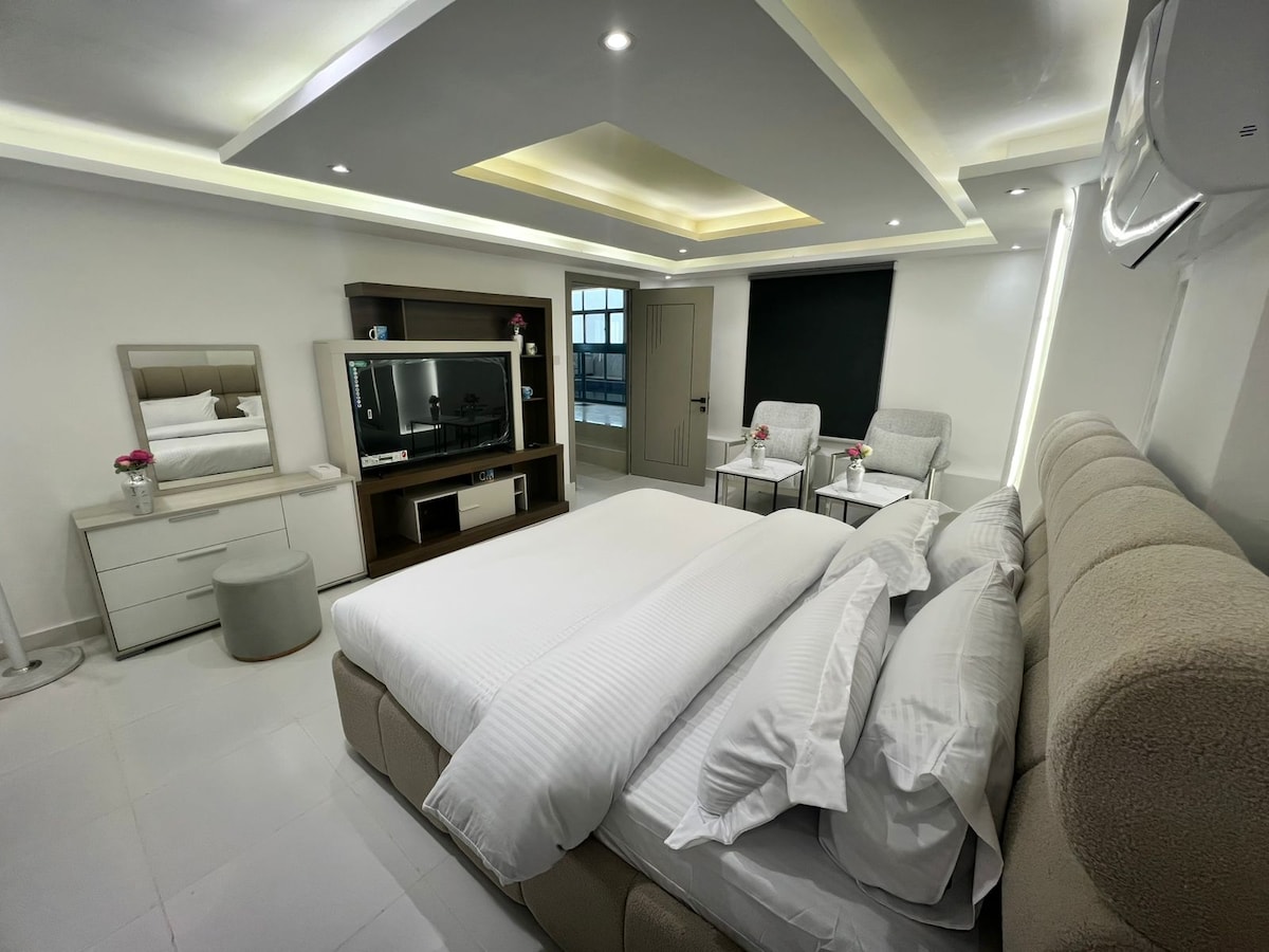 A modern bedroom is displayed, featuring a spacious bed dressed in white linens. Two armchairs with a small table are positioned nearby, and a large television unit is mounted on the wall. Natural light enters through a window, contributing to the bright atmosphere.