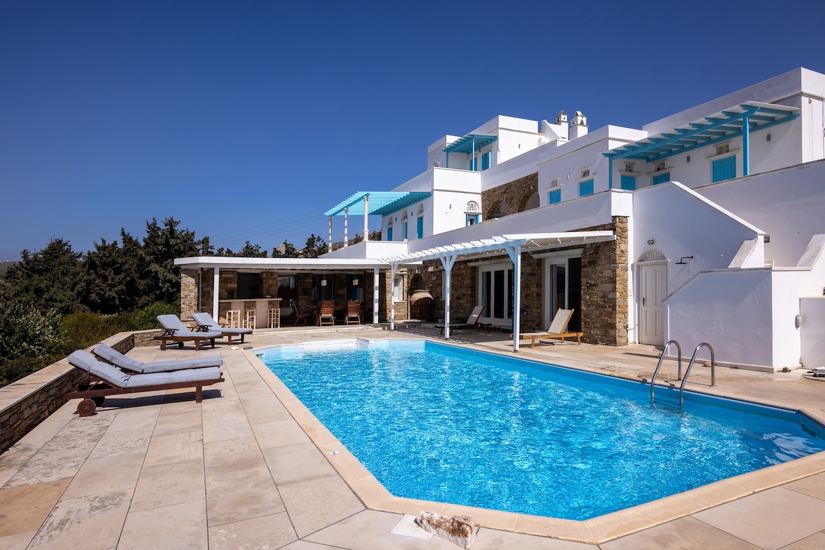 A spacious outdoor area features a large swimming pool surrounded by sunbeds and a stone patio. The villa, characterized by its white and blue architecture, offers sheltered seating and panoramic views of the surrounding landscape underneath a clear blue sky.