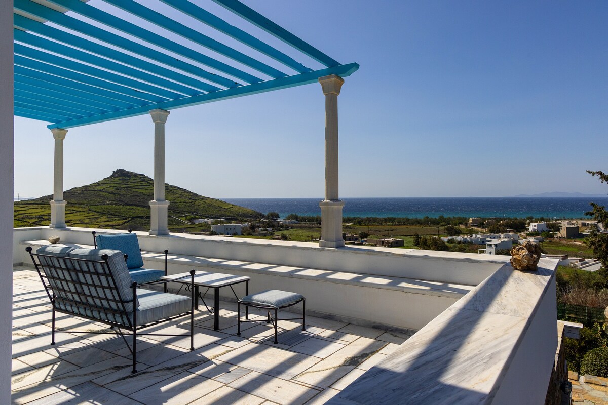A rooftop terrace features a striking blue pergola, offering unobstructed views of the sea and surrounding landscape. Comfortable seating arrangements are placed on a white marble surface, creating a serene space for relaxation. The horizon showcases a blend of blue water and greenery.