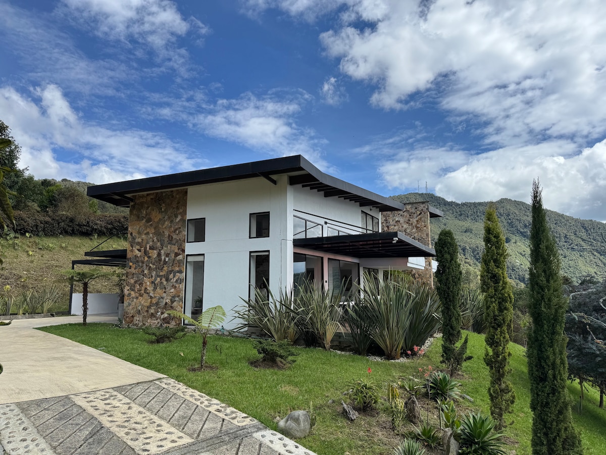 The exterior of the house is showcased with its modern design featuring a mix of stone and white walls. Surrounding greenery and well-maintained landscaping add to the natural setting. The mountains are visible in the background under a partly cloudy sky.
