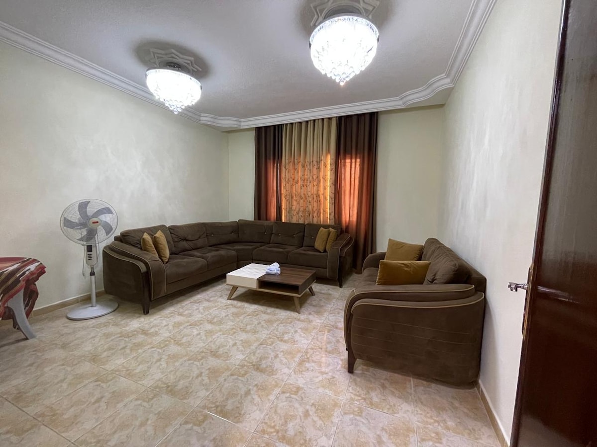 A spacious living area features a large, U-shaped brown sofa, complemented by decorative cushions. Natural light filters through draped curtains, illuminating the textured walls. A small coffee table sits in the center, while a ceiling fan offers a practical cooling option.