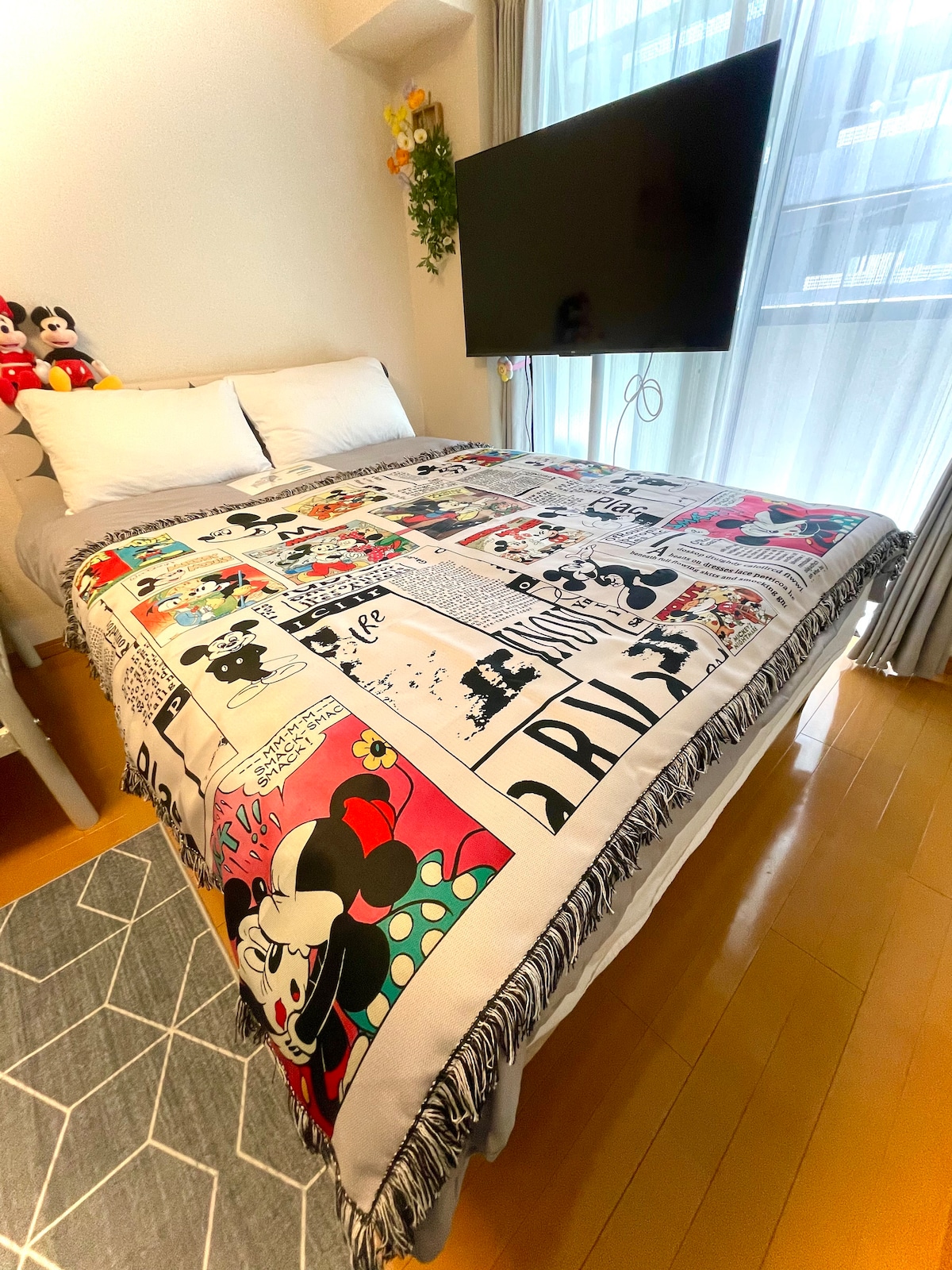 A colorful bedspread featuring a comic strip design is draped over a spacious bed. A large flat-screen television is mounted on the wall at the foot of the bed, and natural light filters through sheer curtains, creating a bright atmosphere.