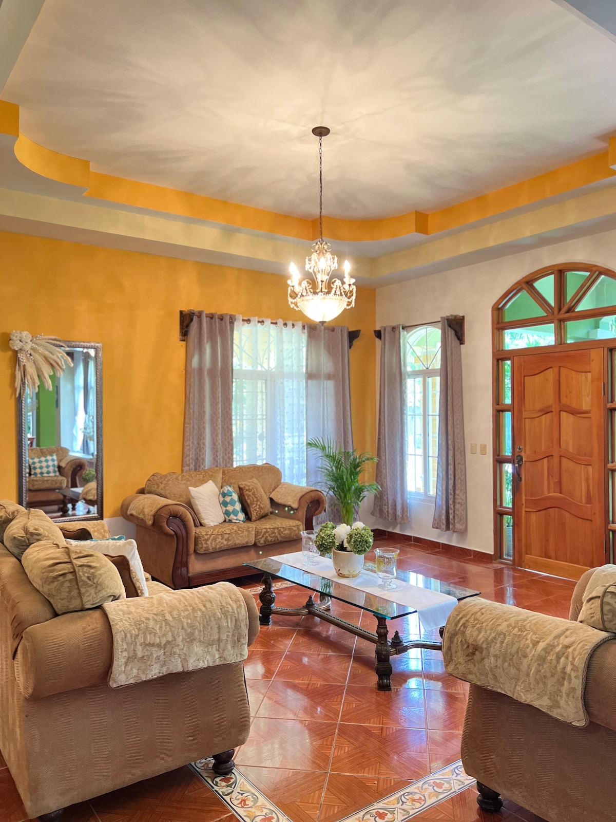 A living room features two cozy sofas facing each other, arranged around a glass coffee table. Large windows are framed with light curtains, allowing natural light to brighten the space. A decorative chandelier hangs from the ceiling, complementing the warm yellow walls.