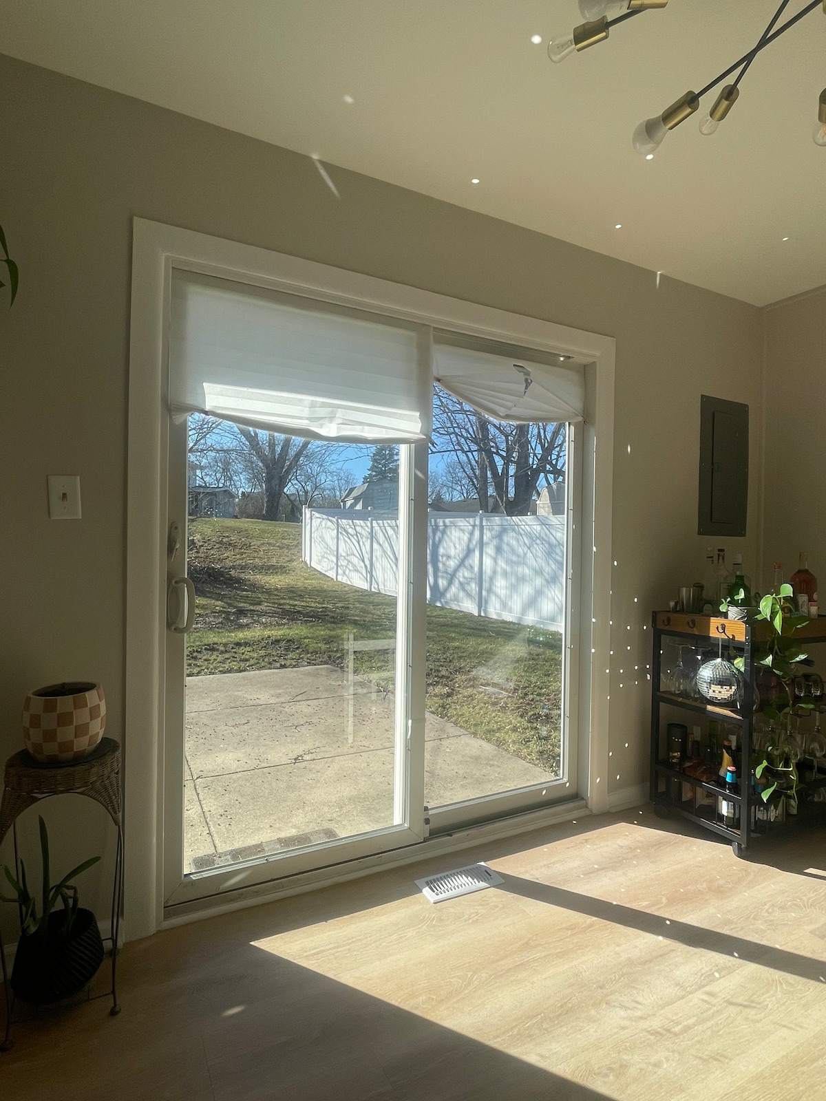 A large glass sliding door allows natural light to enter the room, with views of the backyard and a white fence in the distance. A small bar cart stands nearby, and sunlight creates patterned reflections on the floor.