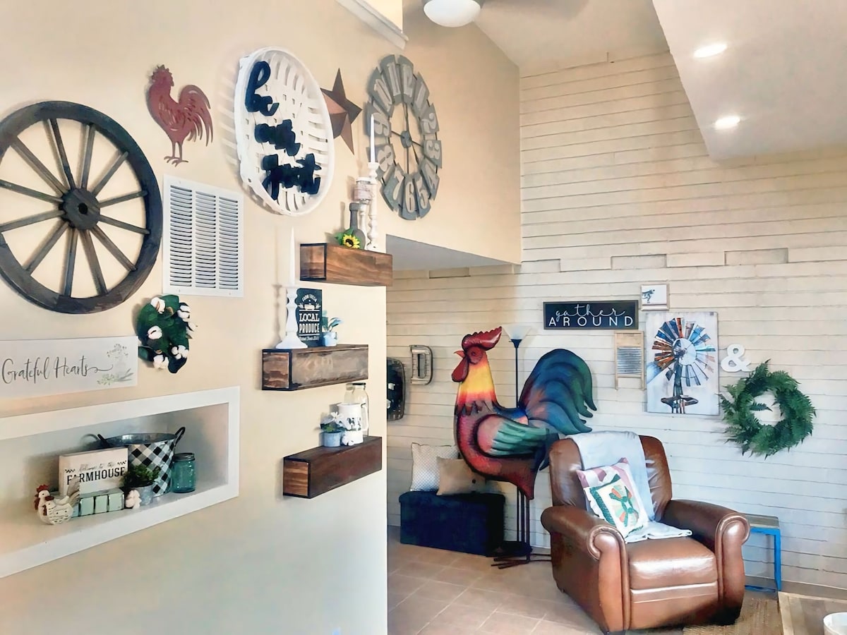 A charming living area features a blend of farmhouse decor, including wooden shelves holding decorative items and a vibrant rooster mural. A comfortable armchair is positioned next to the wall adorned with various framed artworks, enhancing the space's inviting character.