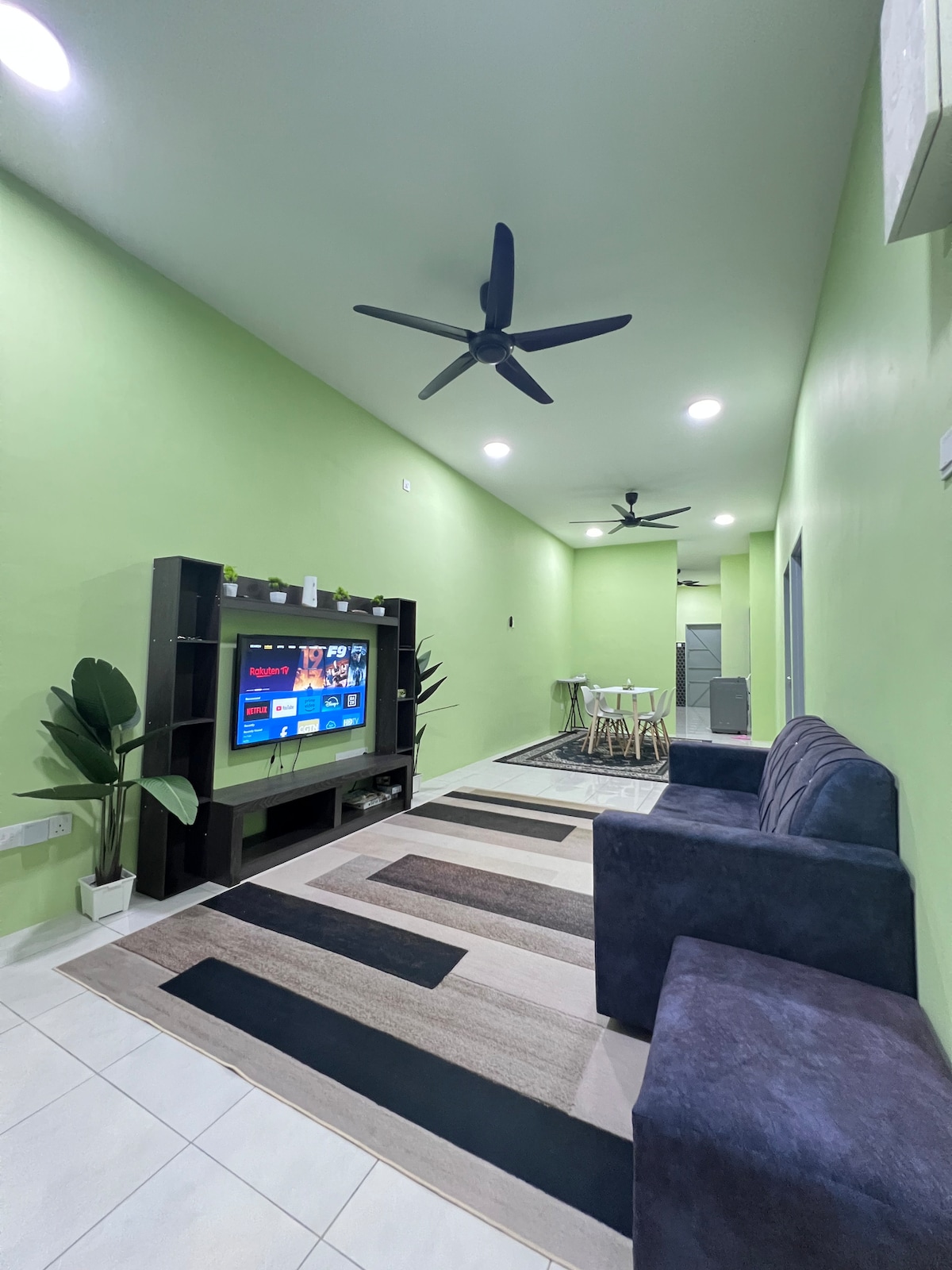 A contemporary living space is presented with light green walls and a cozy dark sofa. A black TV unit houses a flat-screen television, while a small dining area with chairs is visible in the background. A patterned rug adds texture to the tiled floor.