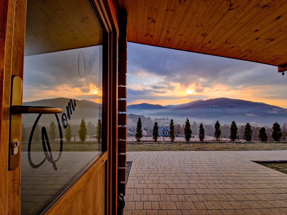 A wooden door partially open reveals a serene landscape, showcasing rolling hills under a sunrise. The sky displays soft pastel hues, while a line of evergreen trees stretches across the horizon, contributing to the tranquil setting.