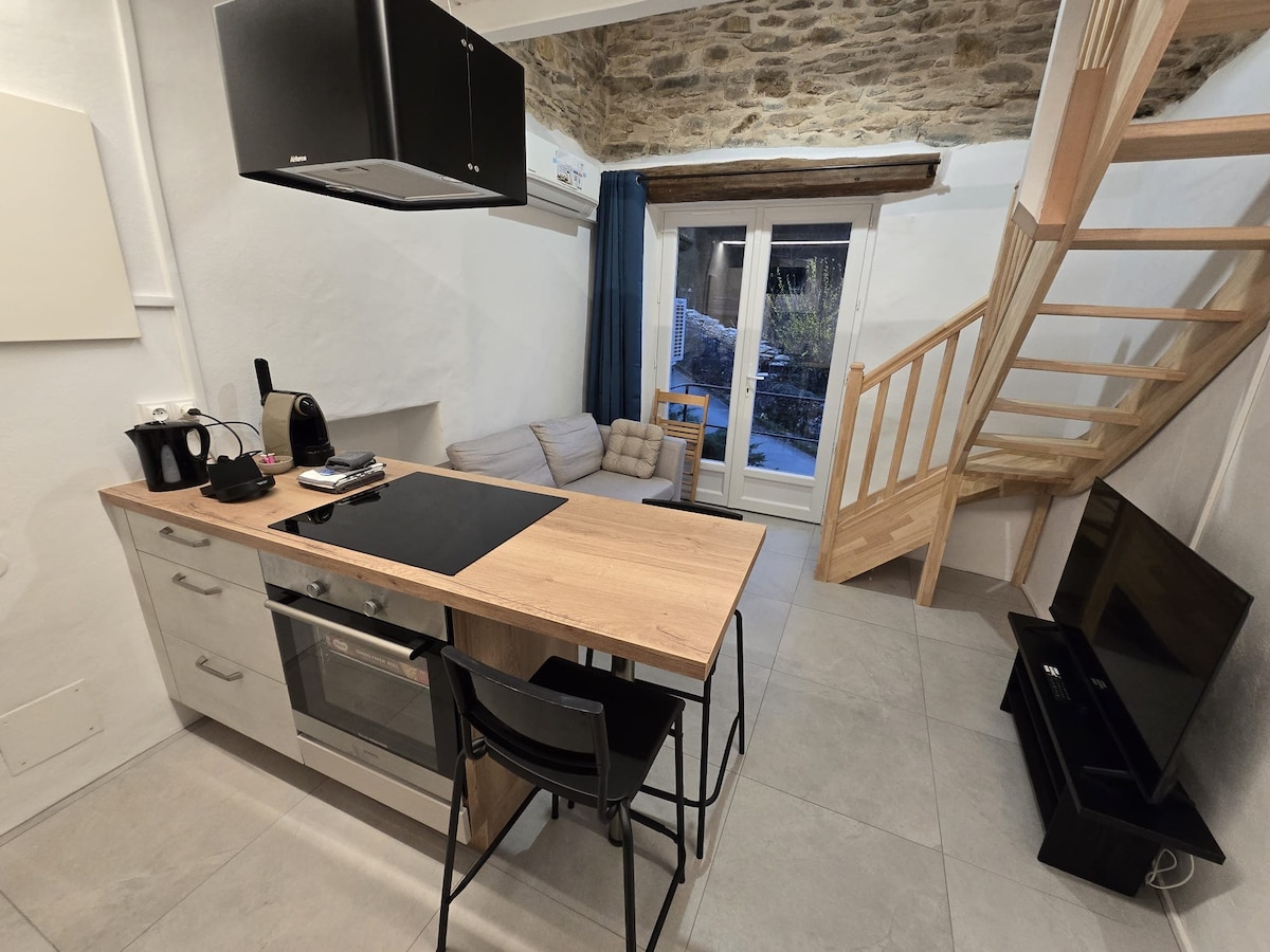 The open-plan living area features a kitchen island with seating and modern appliances, including a stove and coffee maker. A comfortable sofa is positioned near large windows that provide natural light, while a wooden staircase leads to the upper level.