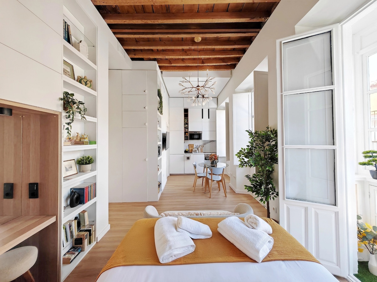 A bright and airy space features a comfortable bed covered in a white duvet and a mustard-colored throw. Open shelves display plants and books. Natural light streams in through double doors leading to a balcony, while a dining area with a table for four is visible in the background.