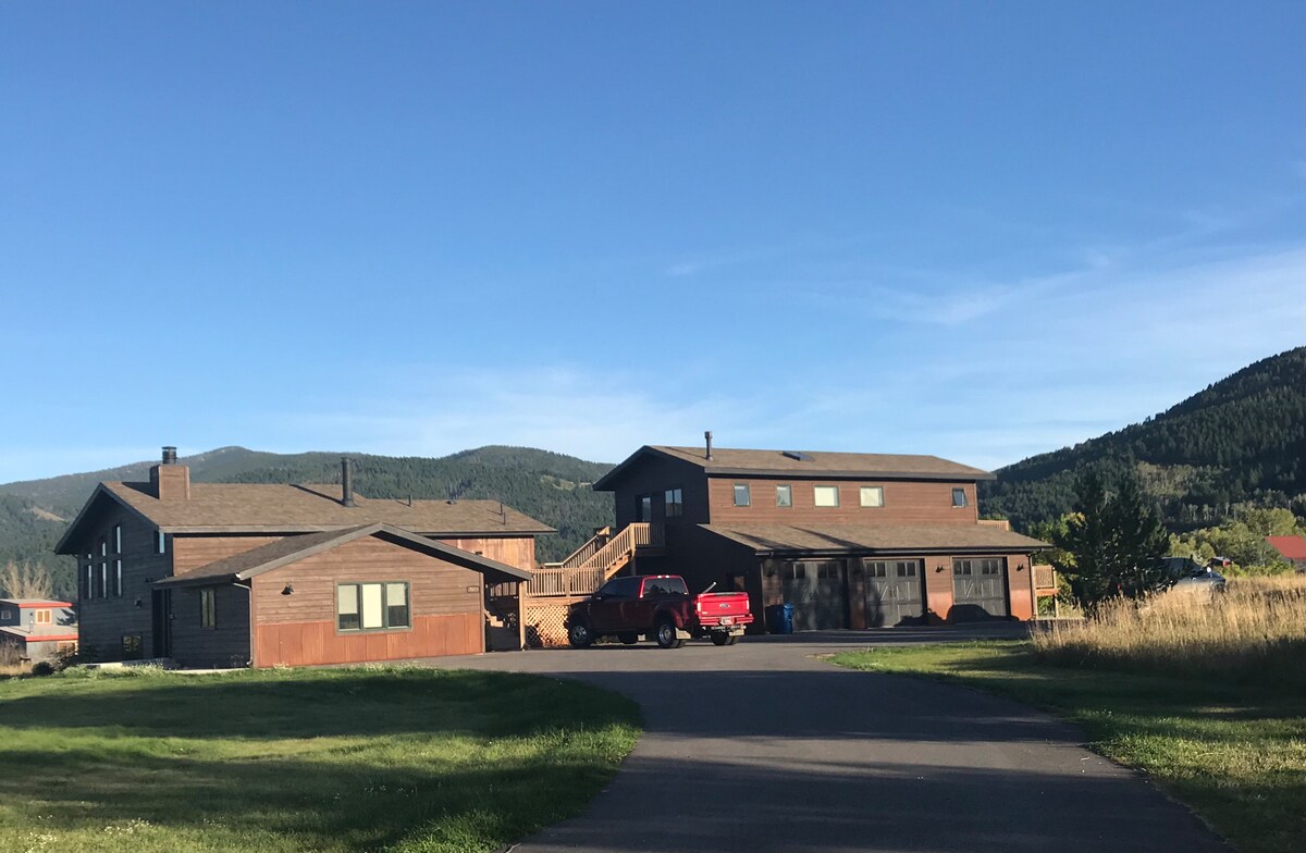 A spacious property featuring multiple buildings, including a two-story loft above a garage. The driveway provides access to the structures, surrounded by open green space and distant mountains under a clear blue sky.