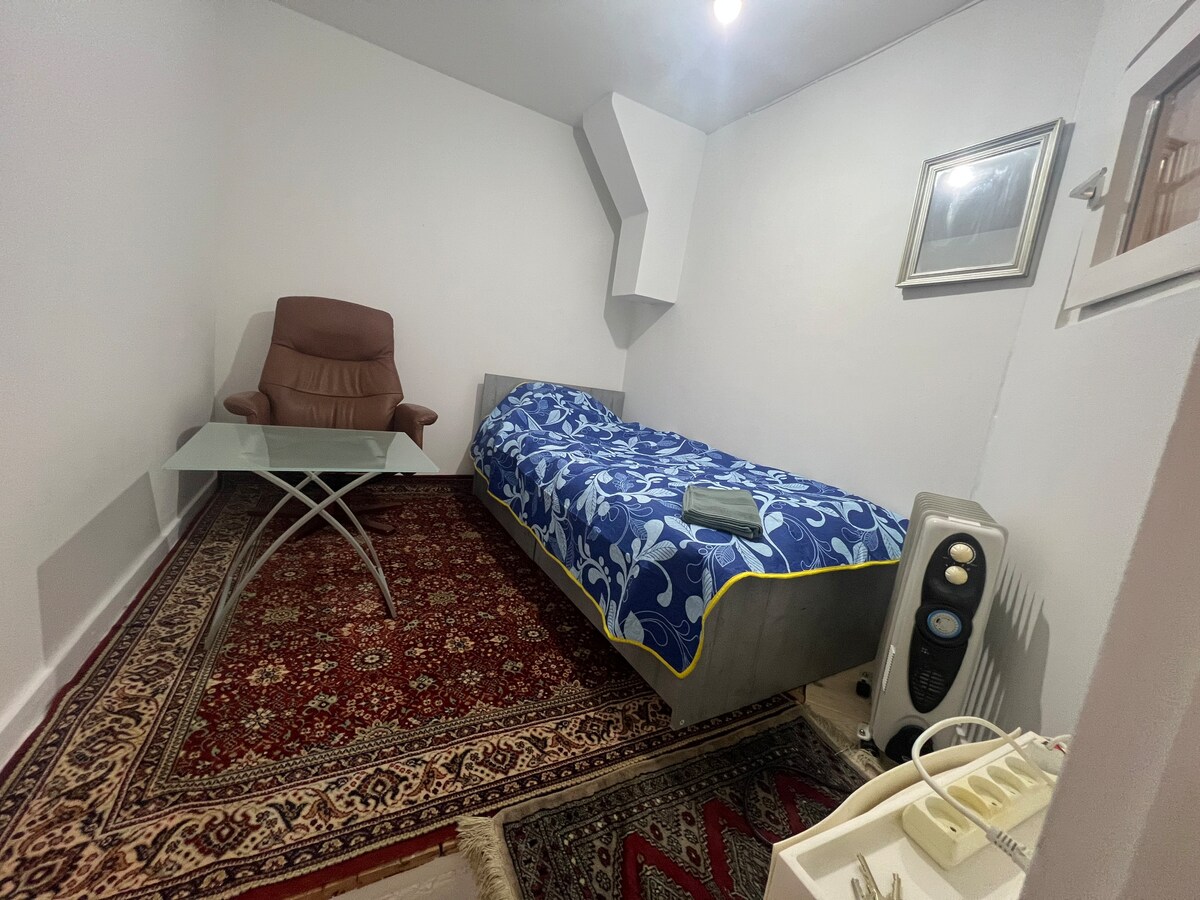 A compact room features a blue floral-patterned bedspread over a spacious bed, complemented by a light brown armchair and a glass table. A decorative rug is positioned beneath the furniture, and a radiator is situated against the wall. Wall-mounted art adds a subtle touch.