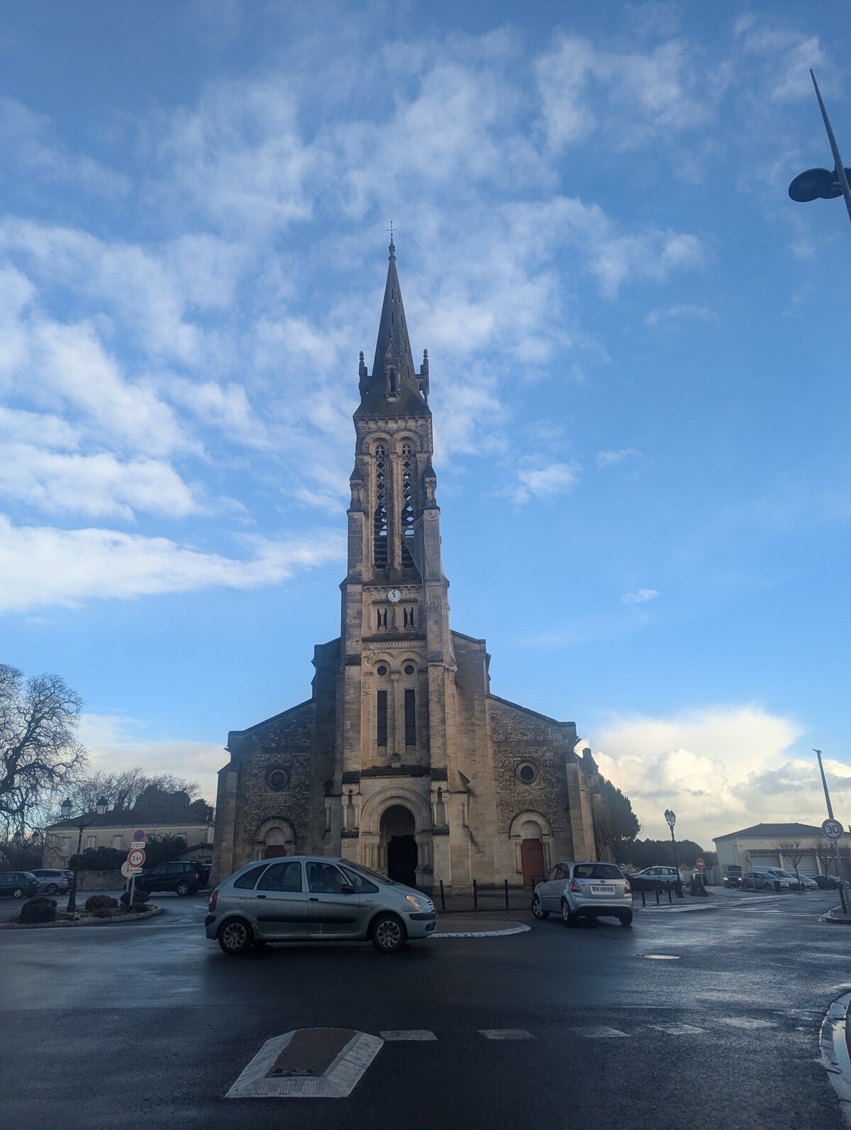 A historic church is depicted with a tall spire reaching towards the sky. The building features intricate stonework and a large entrance, set against a backdrop of blue skies and scattered clouds. Vehicles are parked around the circular intersection in front.