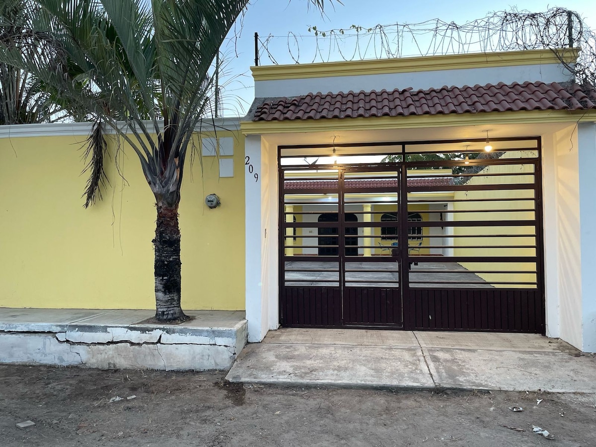 The exterior of a yellow home is presented, featuring a gated entrance with horizontal wooden panels. A palm tree is situated nearby, and a concrete pathway leads to the front door. Barbed wire is visible along the top of the wall, indicating added security.