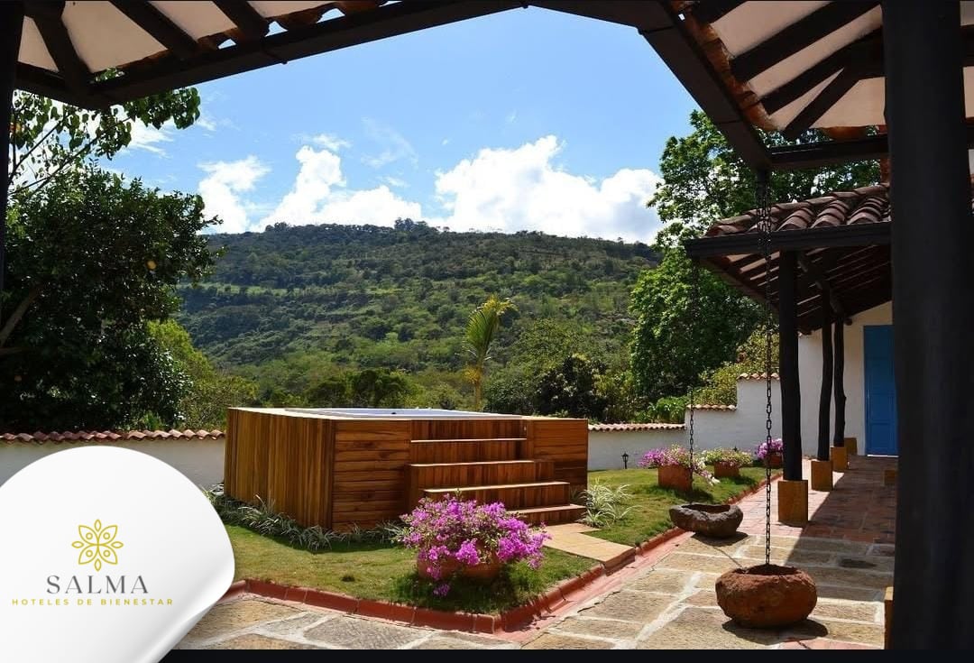 An outdoor terrace showcases a wooden jacuzzi surrounded by vibrant pink flowers and lush greenery. A scenic view of rolling hills under a blue sky is visible in the background, with terracotta roof tiles and a rustic support beam adding charm to the space.