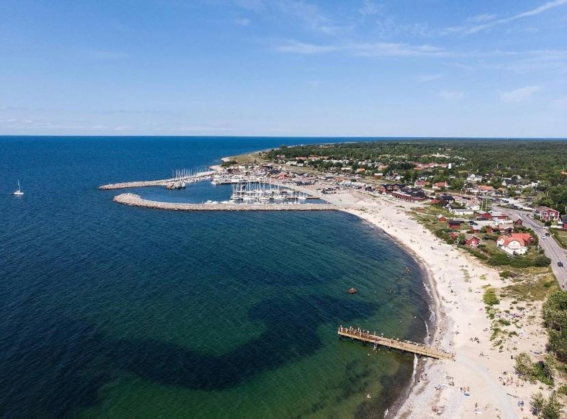 An aerial view of Byxelkrok reveals a curving coastline along the Baltic Sea, with sandy beaches meeting the water. The marina is populated with boats, while a jetty extends out into the sea. Residential and commercial buildings are visible along the shoreline and greenery stretches in the background.