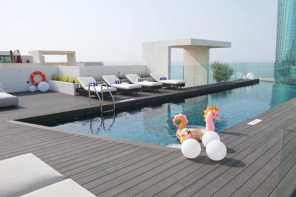 An outdoor pool area is shown, featuring loungers arranged around the pool and floating pool toys. The wooden deck extends around the water, with a city and sea view visible in the background. Sunlight creates a bright yet serene atmosphere.