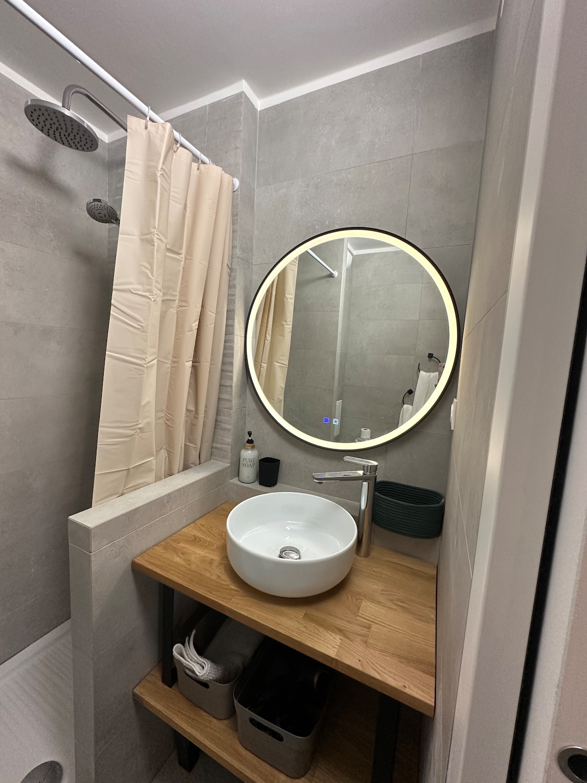 The bathroom features a spacious shower enclosed with a light curtain. A round mirror is illuminated by a soft light above a wooden vanity, which holds a modern sink. Storage baskets are placed below for convenience.