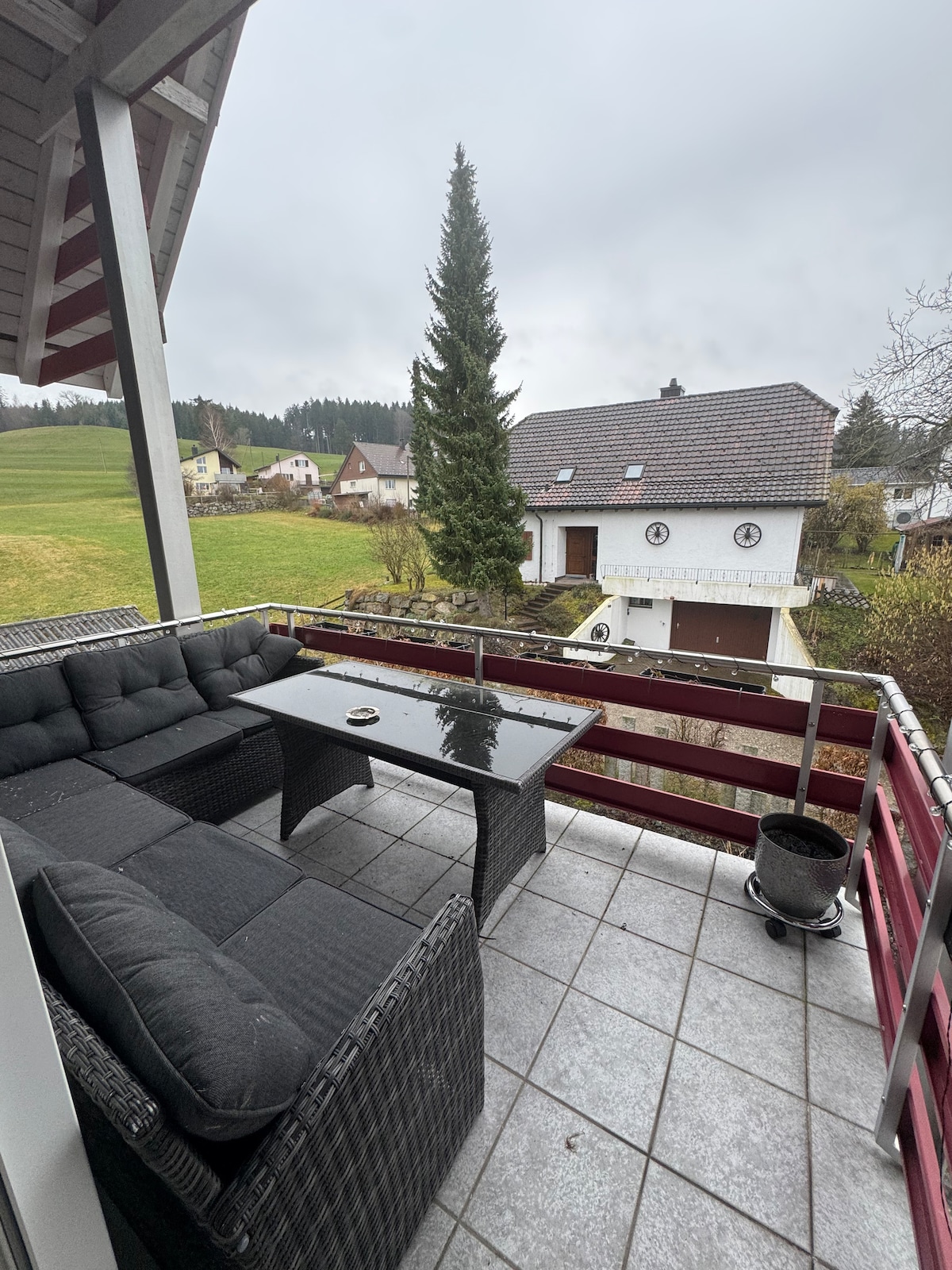A covered balcony features a seating area with dark wicker furniture, including a sectional couch and a glass-topped table. A view of the surrounding greenery and neighboring houses is visible beyond the railing, under an overcast sky.