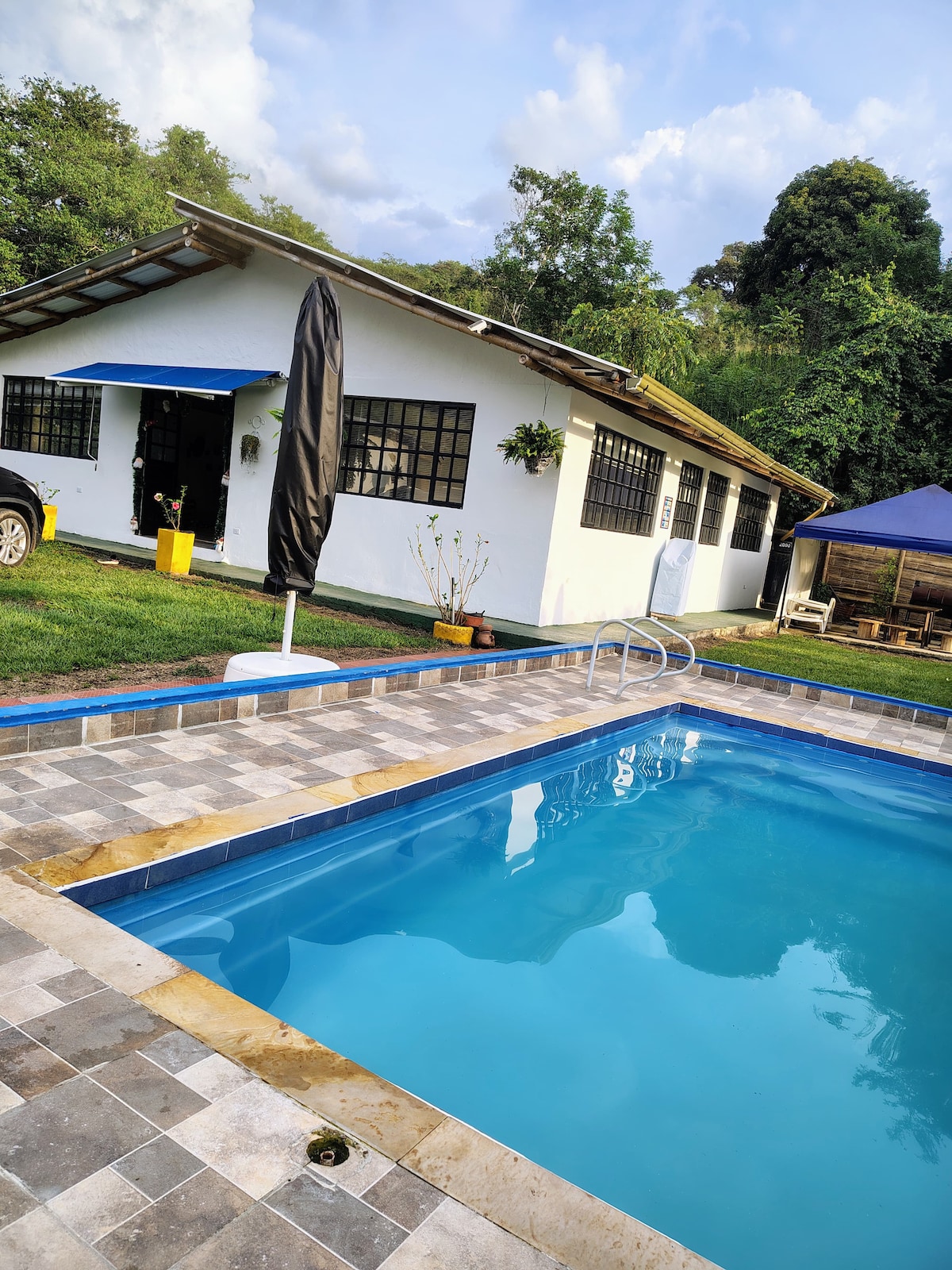 A clear swimming pool is framed by a textured stone patio, with a black umbrella providing shade nearby. A single-story house with large windows is positioned in the background, surrounded by greenery, creating a serene environment.