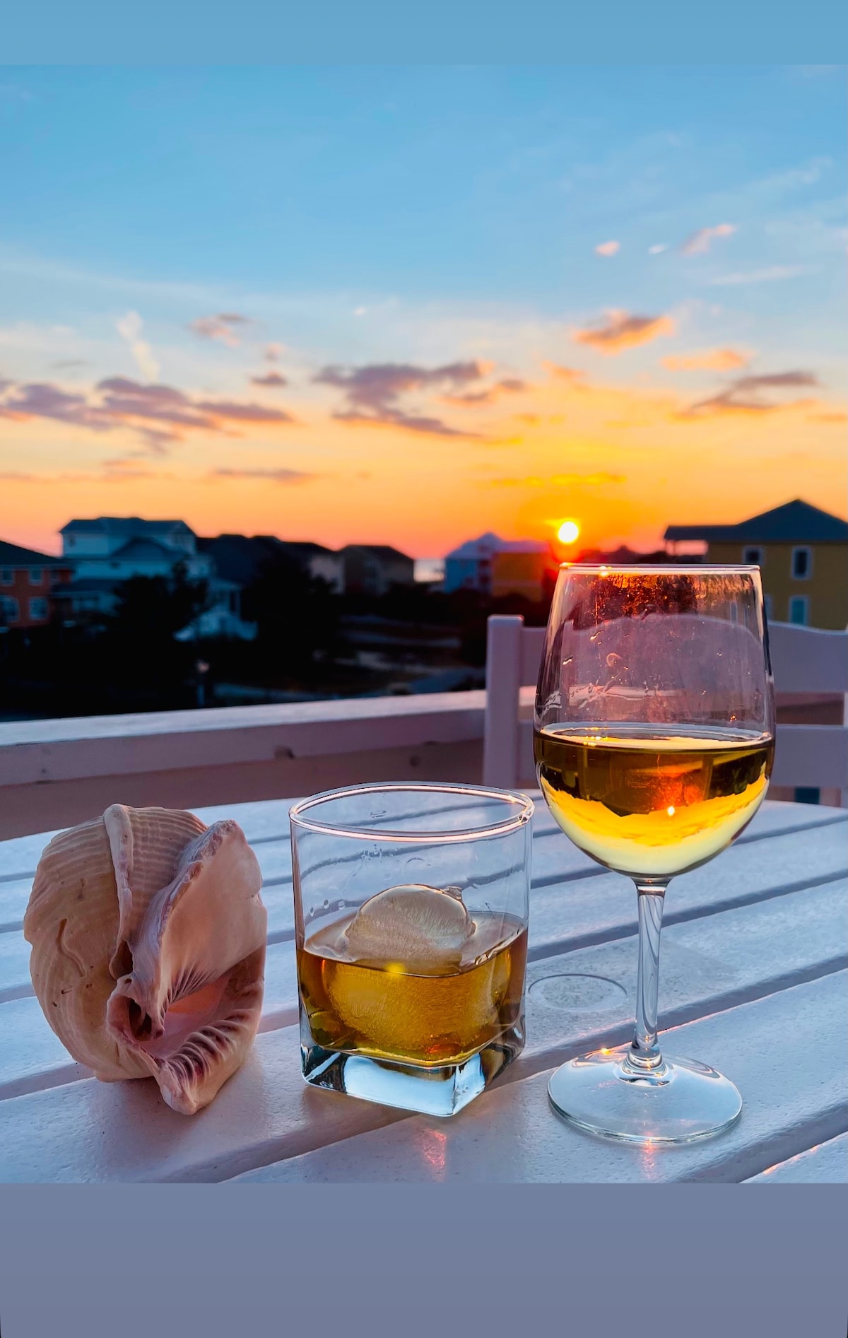 A scenic sunset view showcases a glass of white wine and a lowball glass with a drink, accompanied by a large seashell. The vibrant sky displays hues of orange and pink, while silhouettes of coastal buildings are visible in the background.