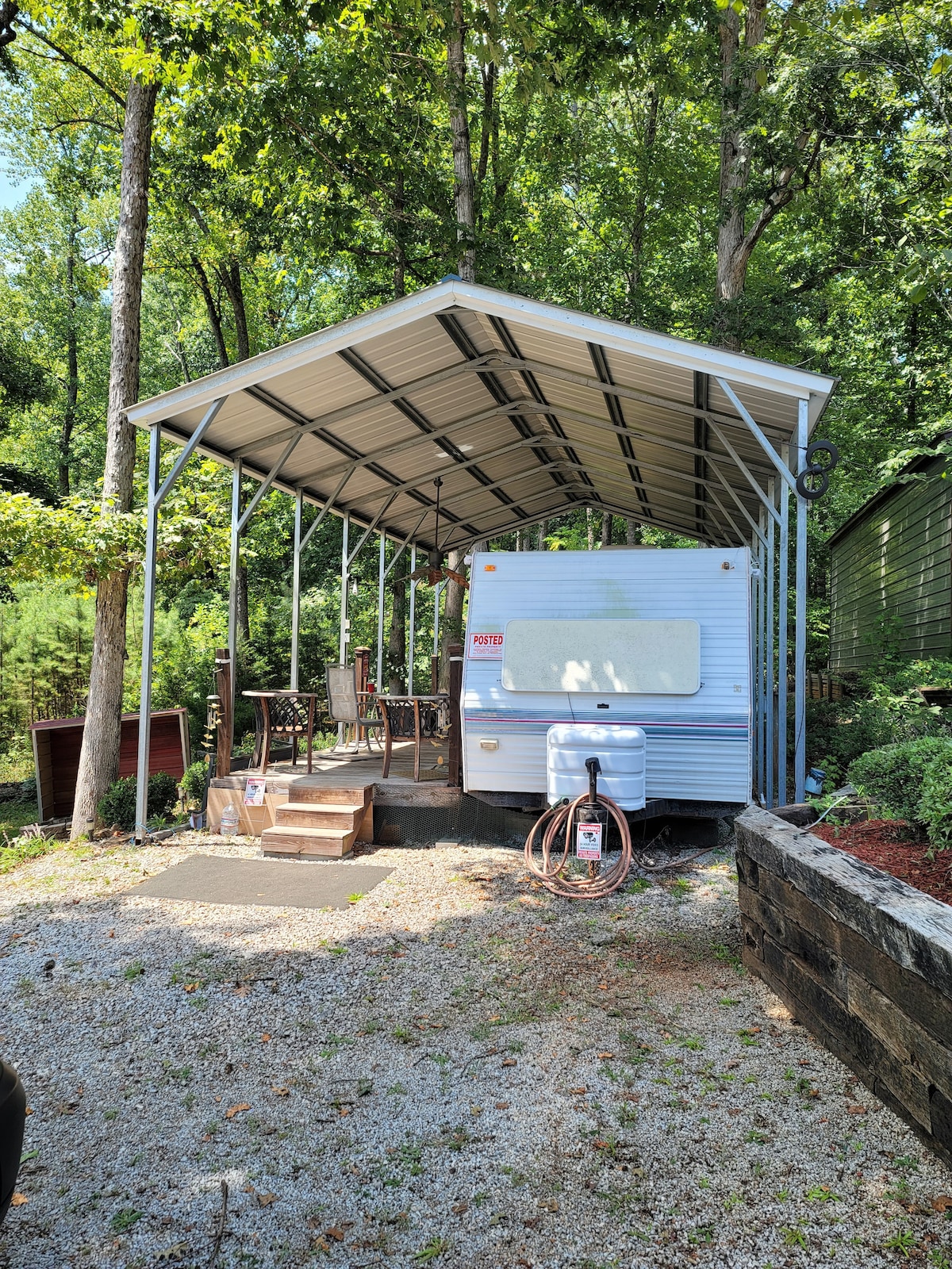 An RV camper is shown beneath a metal awning, providing shelter from the elements. Steps lead to the entrance, with a small outdoor seating arrangement visible nearby. Surrounding greenery enhances the natural setting, contributing to a tranquil atmosphere.