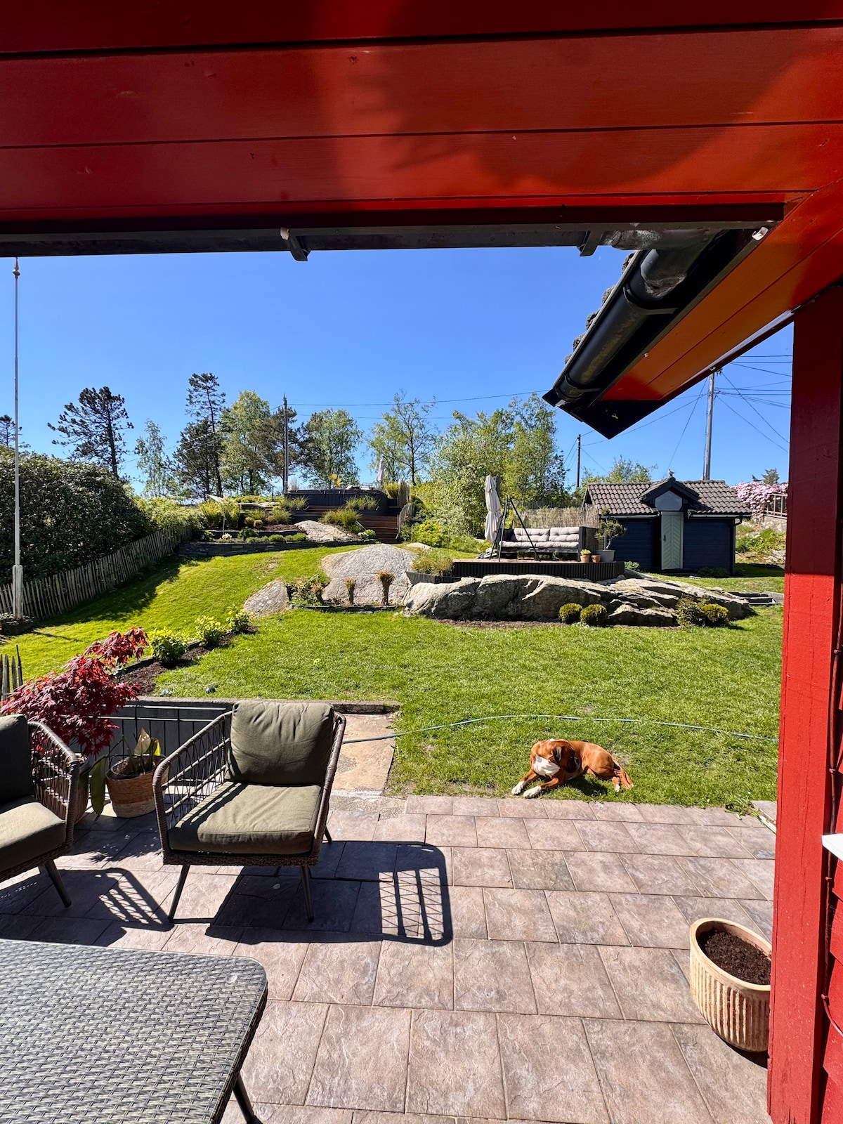 A well-maintained outdoor space features a grassy garden with natural stone elements and surrounding trees. A seating area with two chairs is positioned on a tiled patio, alongside a cozy dog resting on the grass. Beyond, additional garden structures can be seen.
