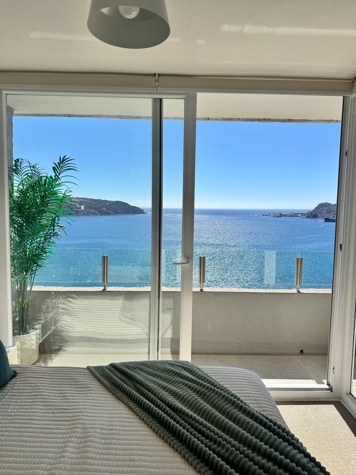 A bright bedroom features floor-to-ceiling sliding glass doors, offering a panoramic view of the ocean. Natural light floods the space, highlighting the serene atmosphere. A potted plant adds a touch of greenery, while a soft blanket is casually draped across the spacious bed.