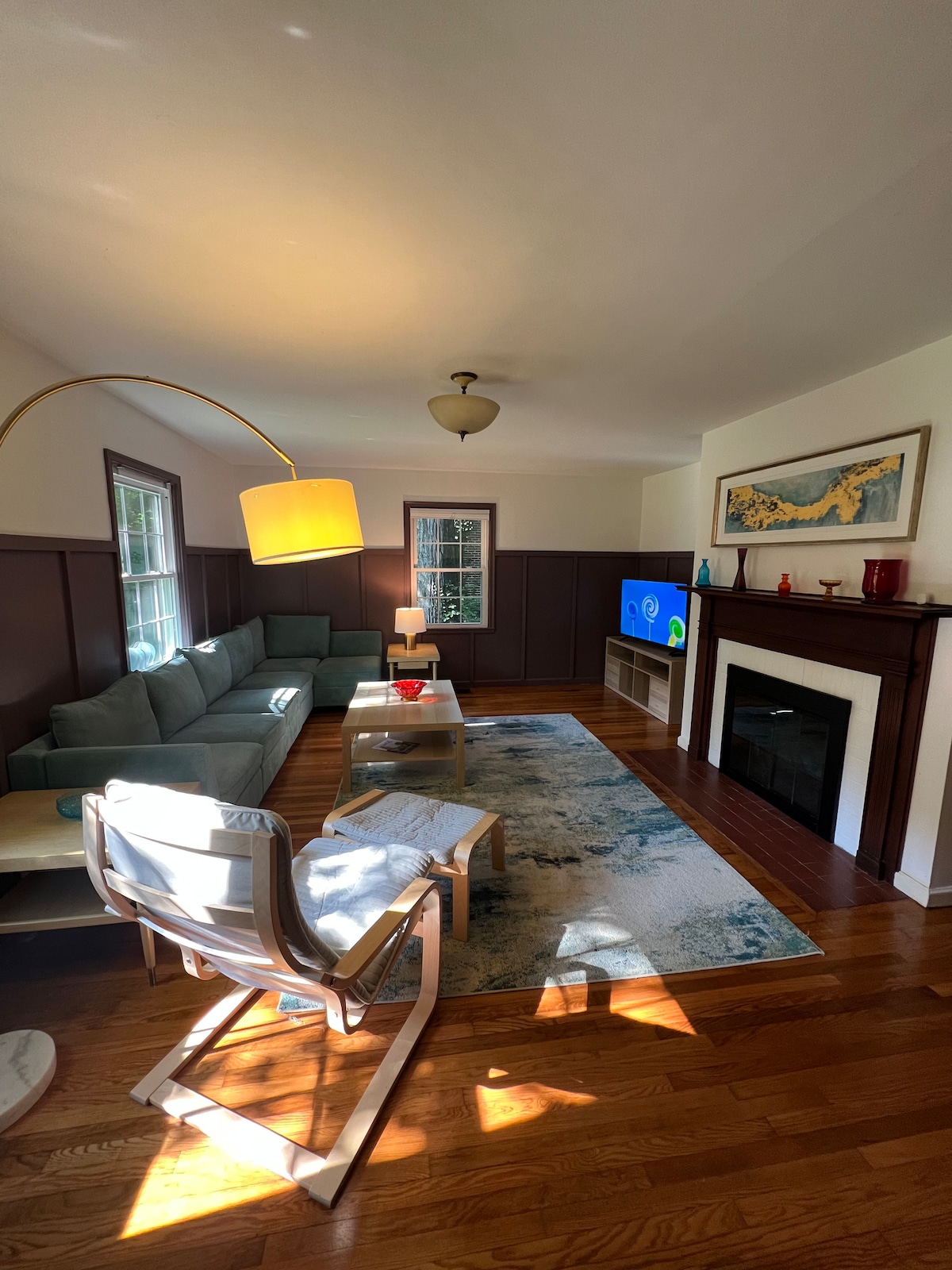 A spacious living area features a large sectional sofa arranged around a central coffee table. A flat-screen TV is positioned on a wooden console. Sunlight filters through the windows, highlighting the warm wood flooring and the decorative area rug beneath.