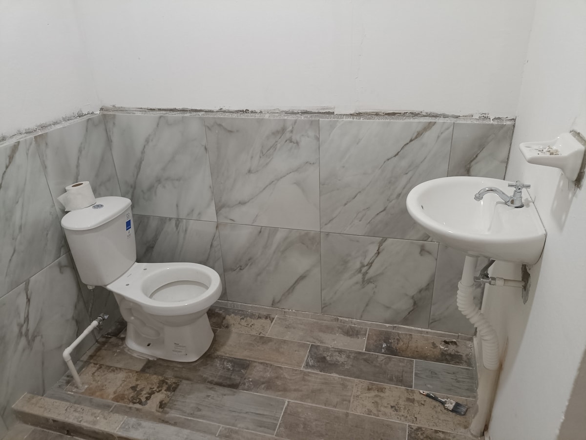 The bathroom features a modern design with marble-style wall tiles and a stylish sink. A toilet is positioned adjacent to the sink, and the flooring is composed of textured tiles. Natural light enters through an opening, enhancing the space.