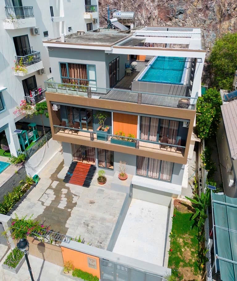An aerial view of a multi-level building is presented, featuring a rooftop pool bordered by a sun deck. The structure showcases multiple balconies with sliding glass doors, surrounded by greenery and landscaped areas. A spacious outdoor area with seating is also visible.