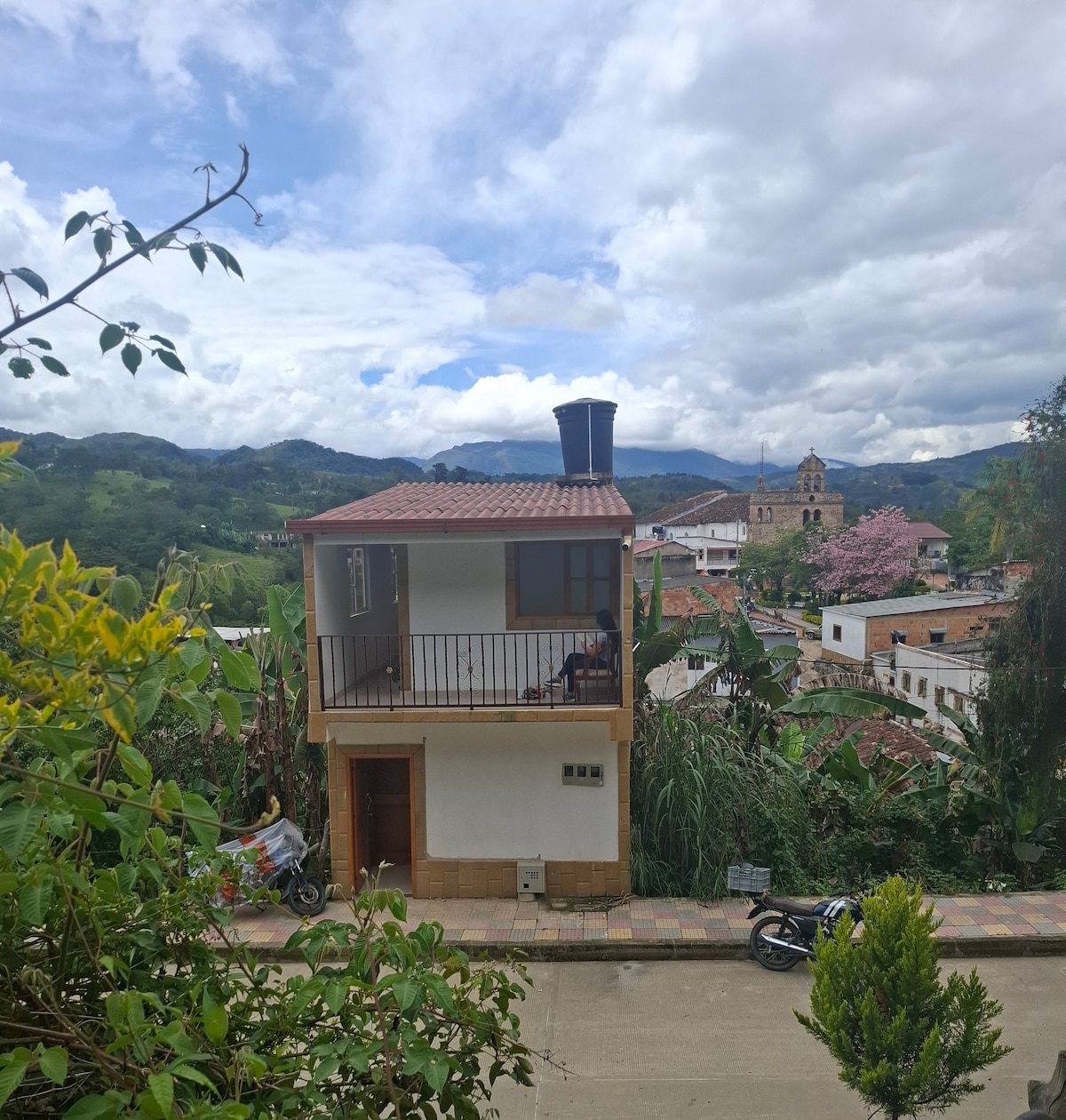 A two-story building is depicted, featuring a terrace with a balcony overhanging the street. The exterior is finished with white and wood accents, and the surrounding landscape includes green hills and blooming trees, along with a historic building visible in the background.