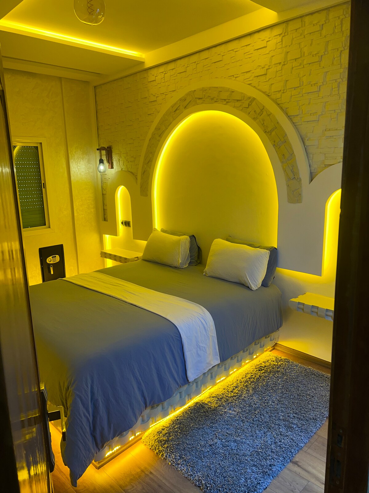 A cozy sleeping area features a spacious bed with soft linens, illuminated by warm yellow LED lights. Soft textures from a plush carpet and the unique archway design on the wall add visual interest. Subtle ambient lighting enhances the modern aesthetic.
