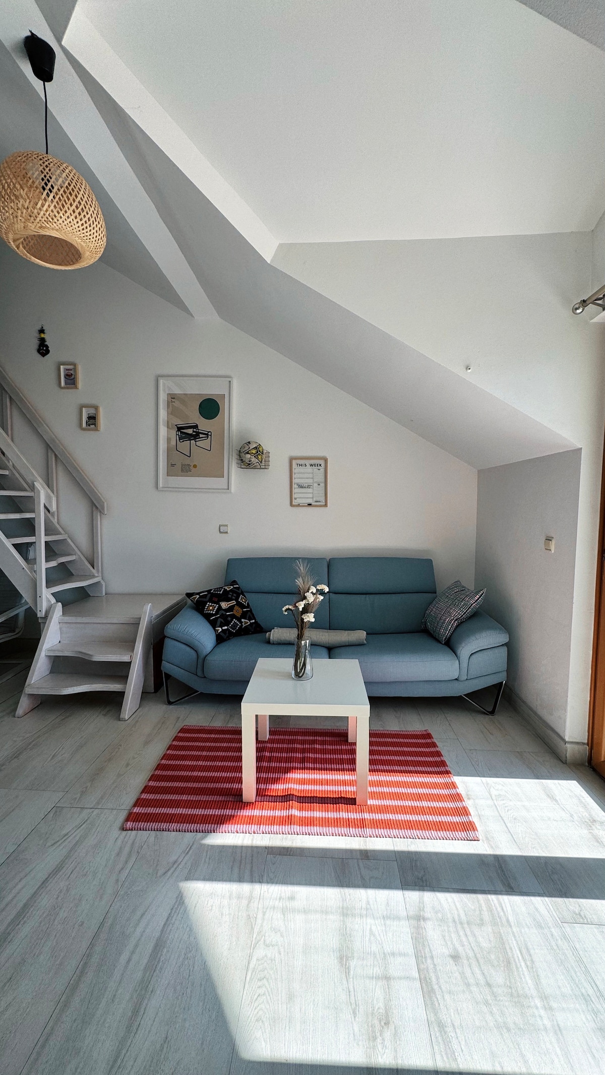 A bright living area is presented with a light blue sofa, adorned with patterned cushions. A small white coffee table sits in front, complemented by a vase of flowers. Sunlight highlights the textured rug in red and white stripes. Stairs lead to an upper level.