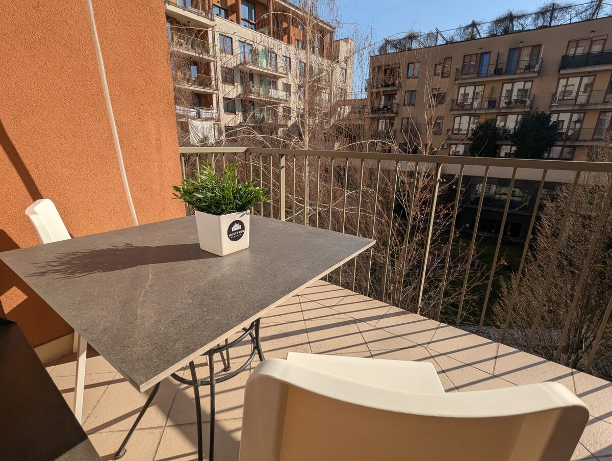 A balcony space is visible with a modern, square table topped with a small plant in a pot. The railing offers an unobstructed view of the surrounding buildings and landscaped area below, allowing for a peaceful outdoor setting.