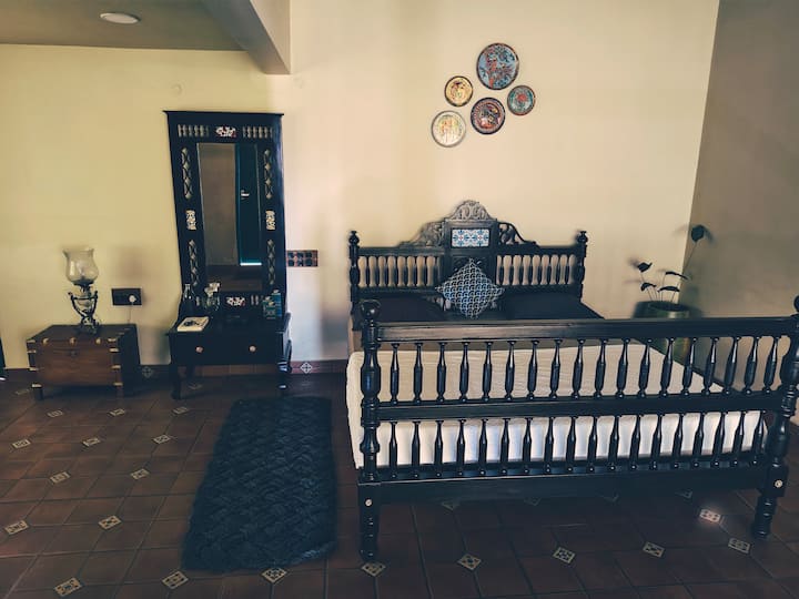 Një dhomë gjumi e stilit kolonial me një krevat druri të errët të gdhendur, komodinë të vjetër dhe pllaka me ton të ngrohtë. Pllakat dekorative të murit shtojnë bukuri, ndërsa ndriçimi i butë dhe mobiliet klasike krijojnë një ambient të rehatshëm, elegant dhe ftues.