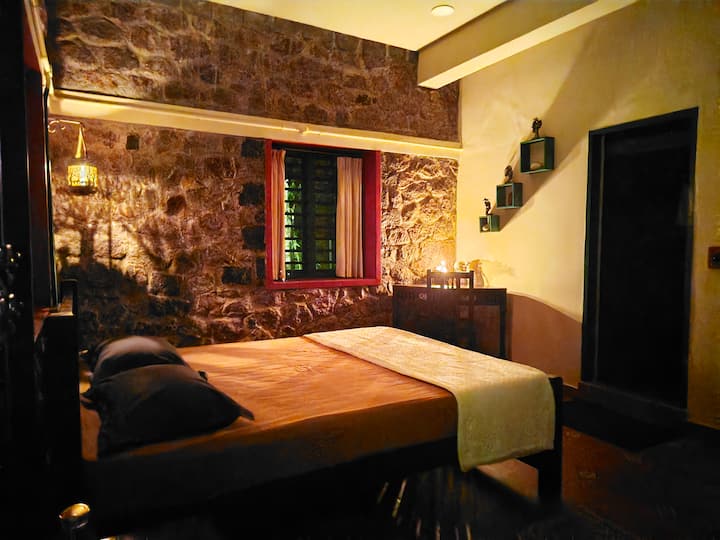 A cozy retreat featuring a queen-size bed, rustic stone walls, and warm lighting. Large windows frame stunning views of tea plantations, meadows, and rolling hills.