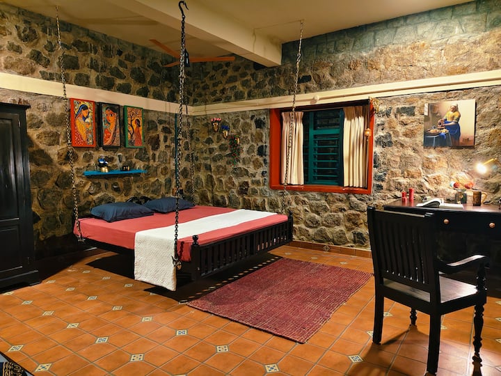 A secluded retreat featuring a king-size hanging bed, rustic stone walls, and warm lighting. Large windows offer sweeping views of meadows and hills, creating a serene escape.
