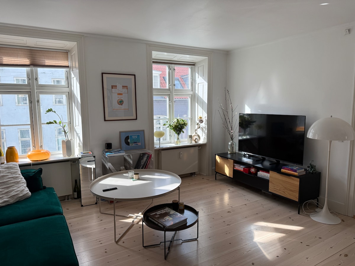 A bright living room is shown, furnished with a large green couch and a modern coffee table. Two windows allow natural light to fill the space, and a TV is set on a sleek black media console with wooden accents. Light-colored wood flooring enhances the airy atmosphere.