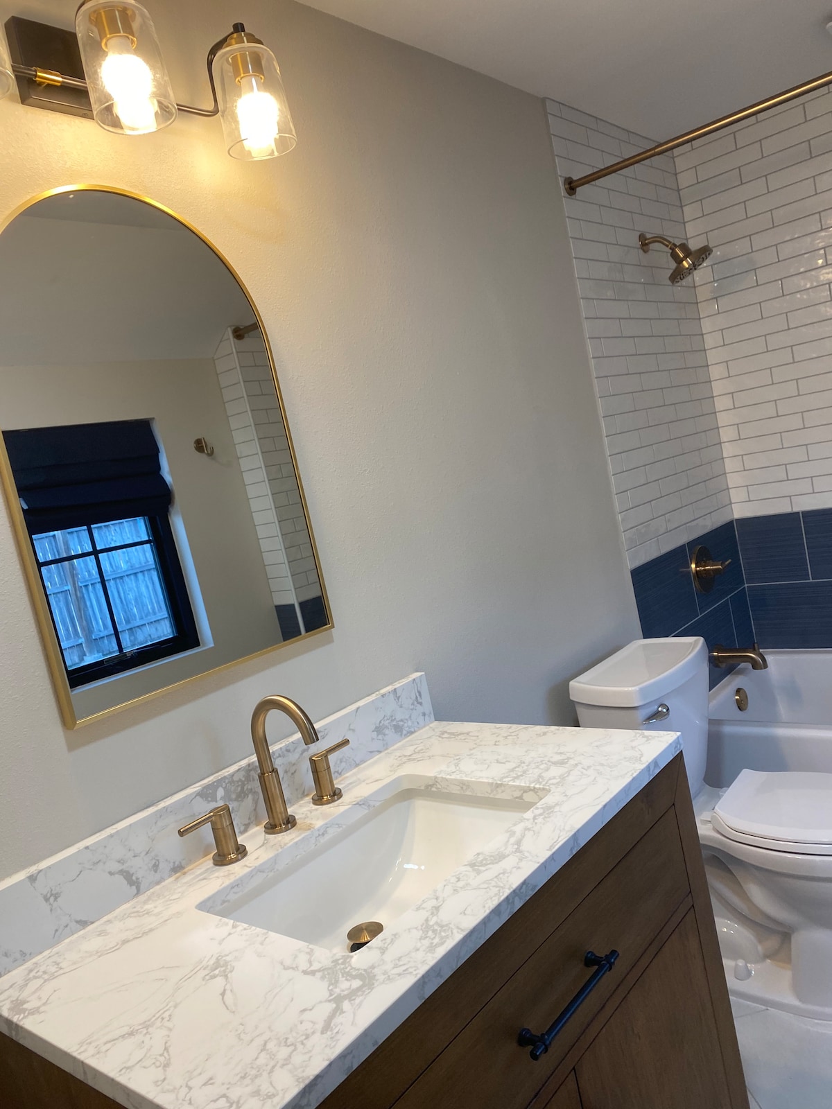 The bathroom features a marble countertop with a modern sink beneath a round mirror. Light fixtures hang above, illuminating the space. A combination of white and blue tiles accentuates the shower area, while a stylish toilet is positioned next to the shower.